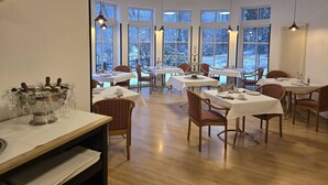 Un espace repas comprenant plusieurs tables et chaises, de grandes fenêtres donnant sur la verdure et un lustre.