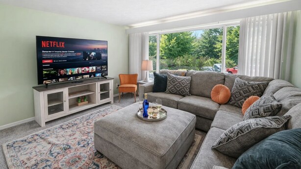 Una sala de estar con un sofá gris, una mesa de centro, un televisor sobre un mueble bajo y una ventana con cortinas.