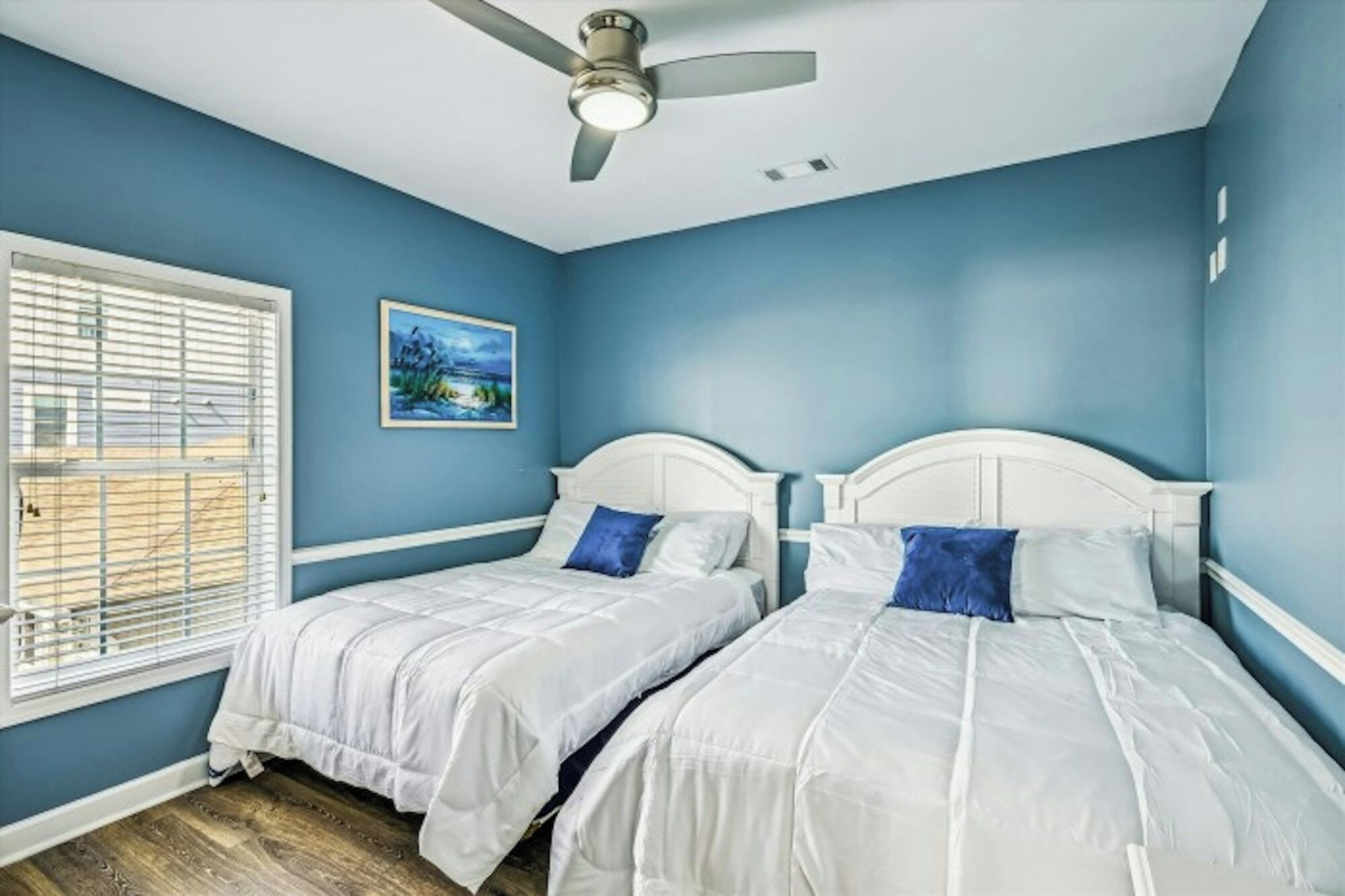 A bedroom with two beds, a ceiling fan, a window with blinds, and a painting on the wall.