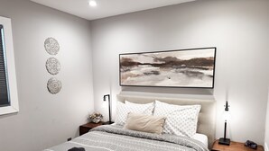 A modern bedroom with a large bed, a headboard, a nightstand, a lamp, and a wall-mounted painting.