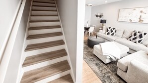 A staircase with wooden steps leading to a landing, a living room with a sofa, ottoman, and a coffee table, and a dining area with a table and chairs.