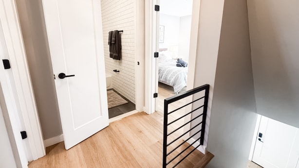 Un escalier avec une rampe en métal noir menant à une chambre à coucher où se trouve un lit recouvert d’une couverture grise.
