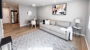 Un salon moderne avec un canapé gris, une table à manger avec des chaises, un coin cuisine avec un réfrigérateur et un tableau accroché au mur.
