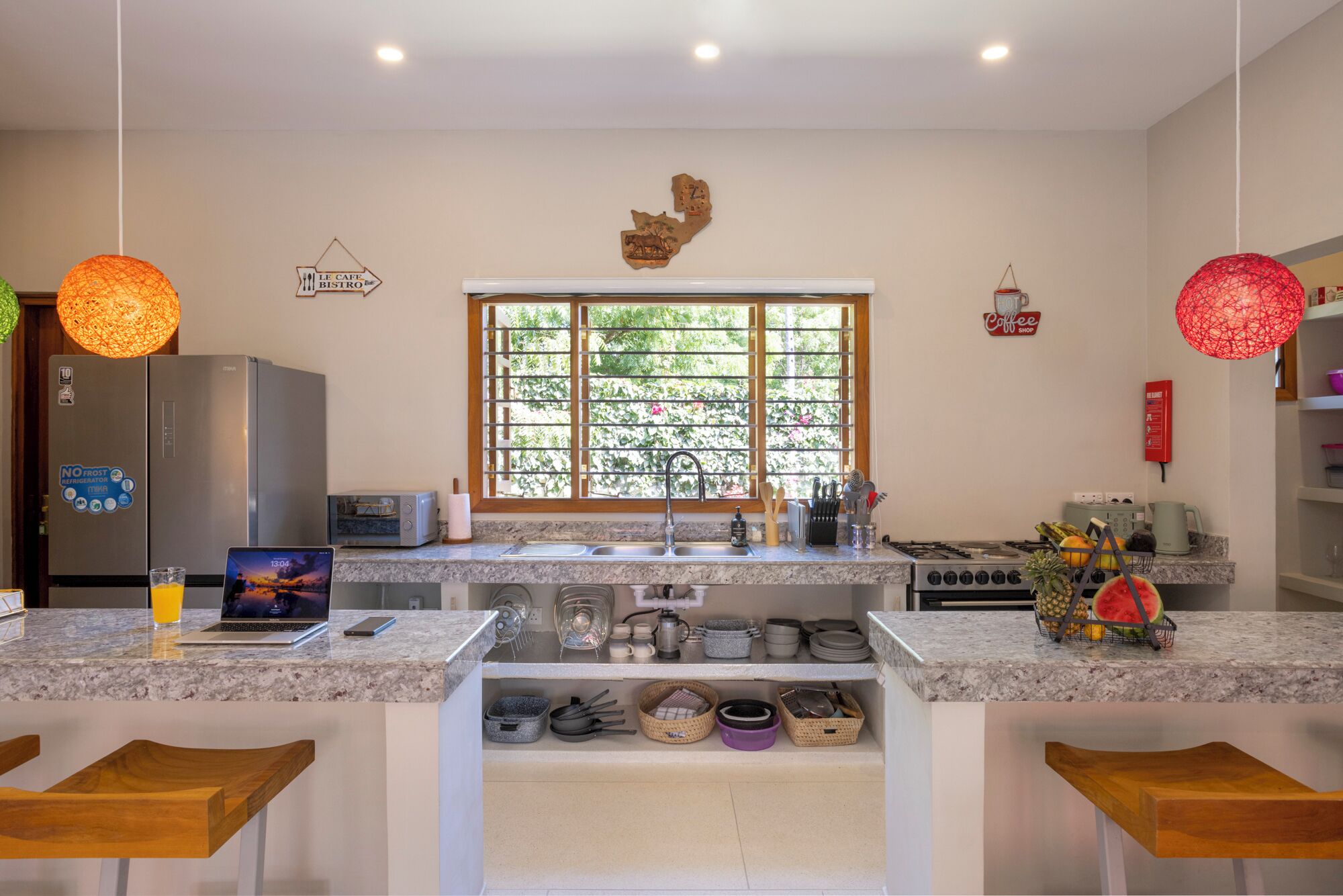 A modern kitchen with granite countertops, a stainless steel refrigerator, a microwave, and a gas stove. There are wooden stools at the counter, a window with a view of greenery, and decorative wall art.