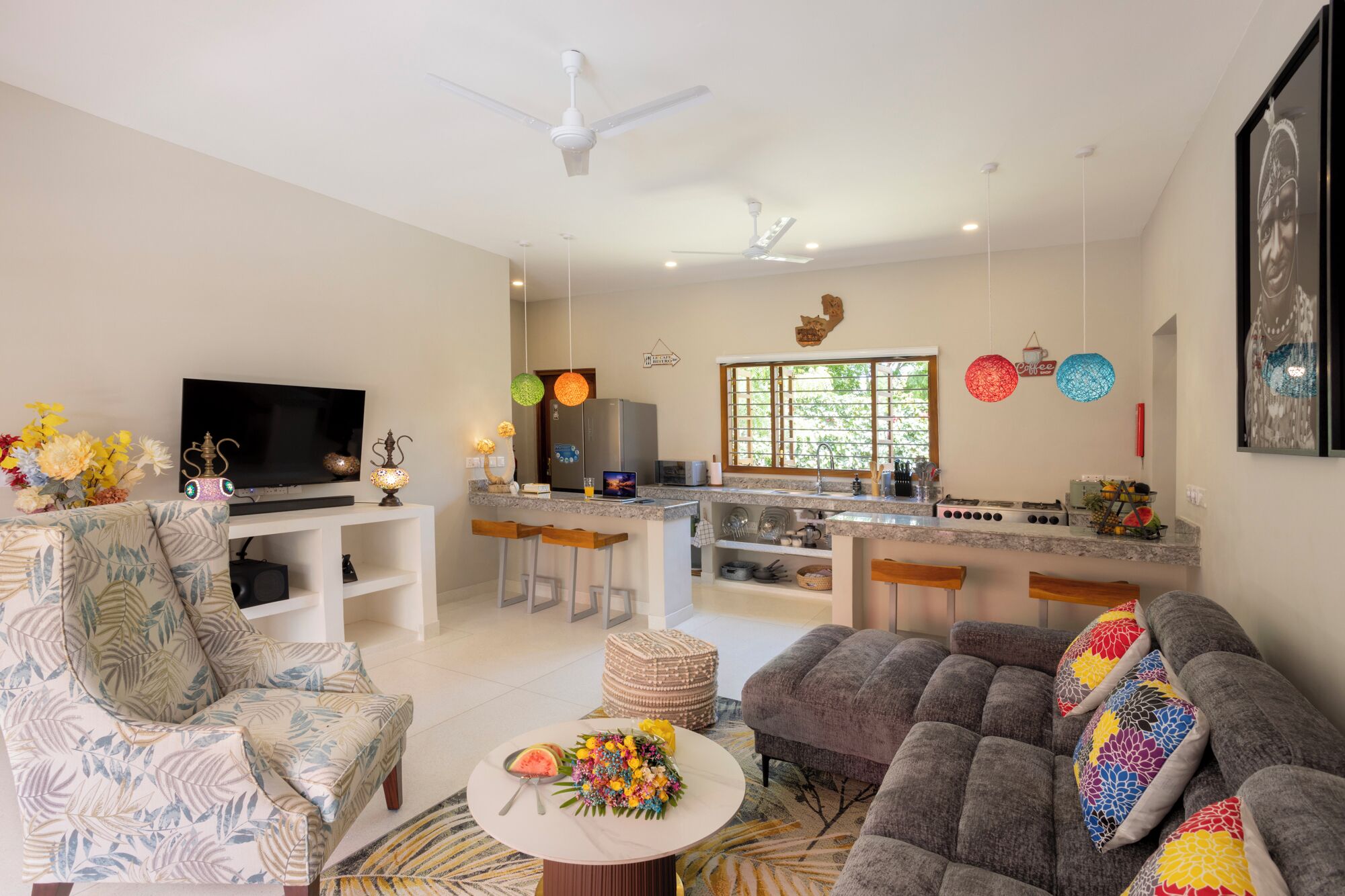 A modern living room with a TV, comfortable seating, a kitchen area with bar stools, and a dining table with a floral centerpiece.