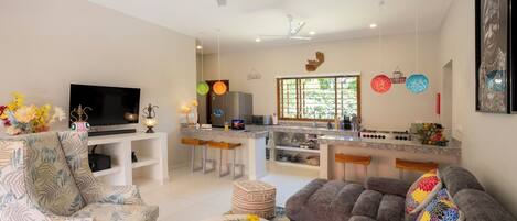 A modern living room with a TV, comfortable seating, a kitchen area with bar stools, and a dining table with a floral centerpiece.