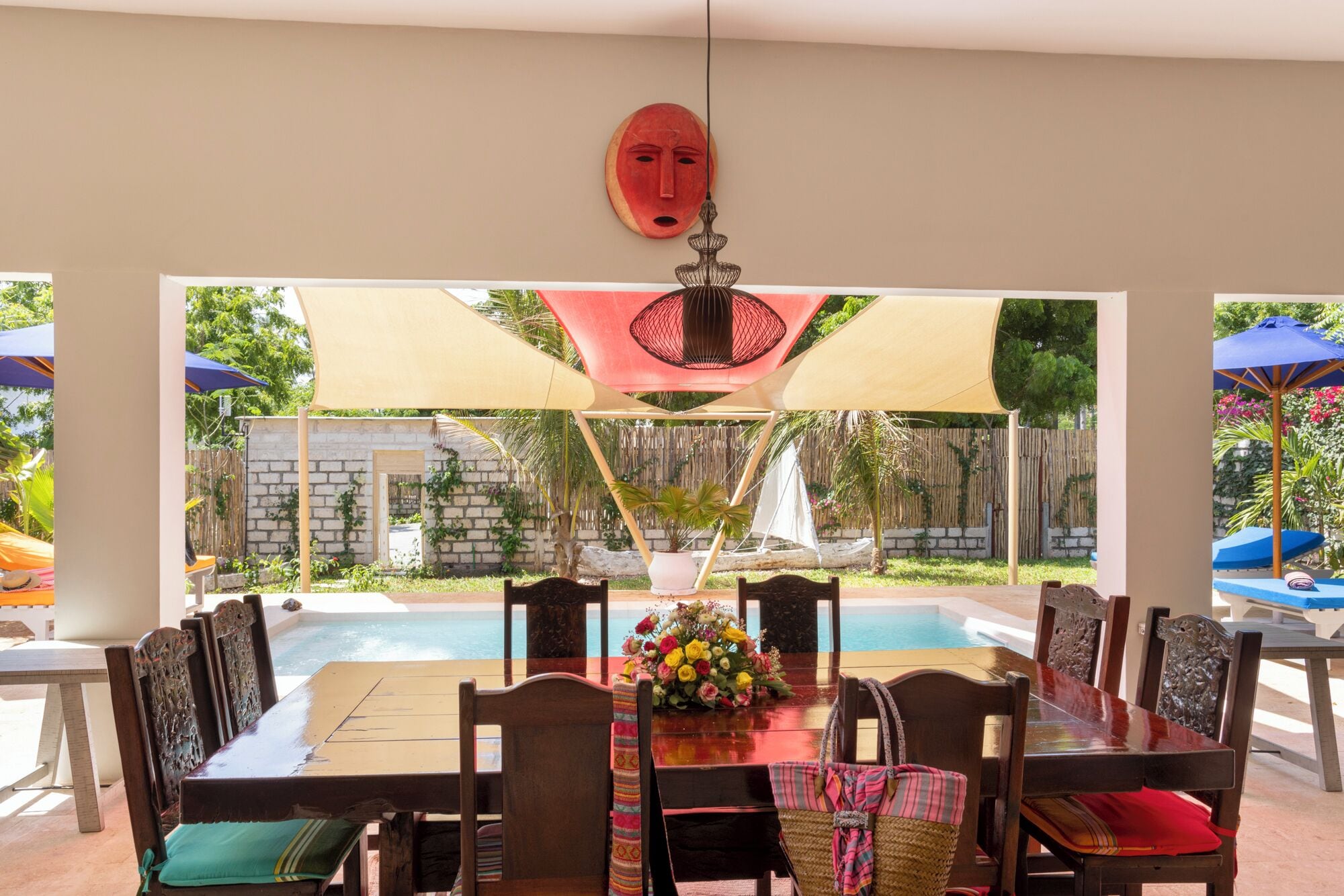 A dining area with a pool view, featuring a table with chairs, a hanging mask, and umbrellas.