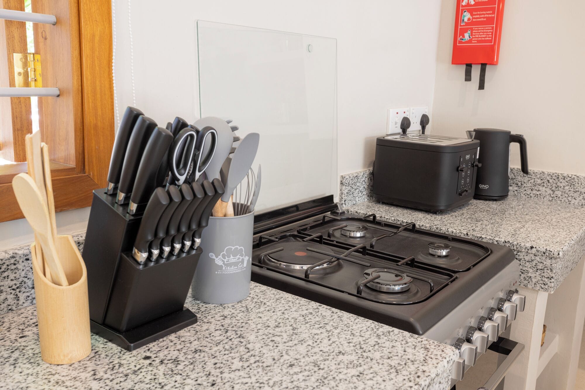 A kitchen with a gas stove, a toaster, and a coffee maker. There is a knife block with knives and a wooden spoon. A fire extinguisher is mounted on the wall.