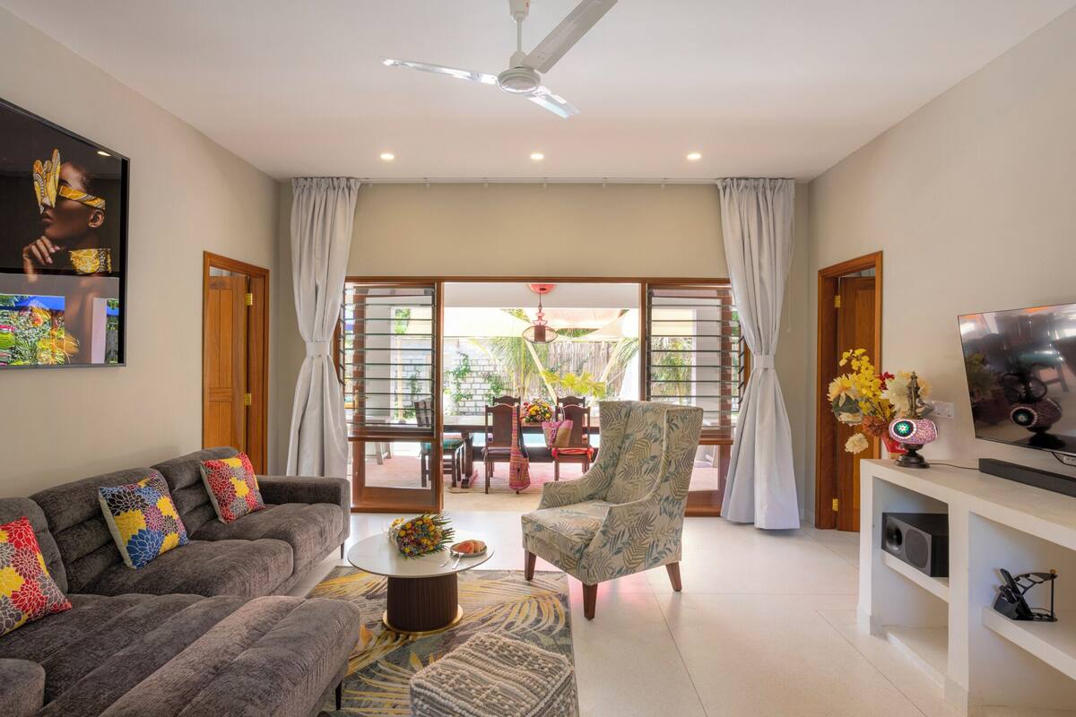 A spacious living room with a grey sofa, patterned armchair, and a coffee table. A television is mounted on the wall, and a ceiling fan is visible.
