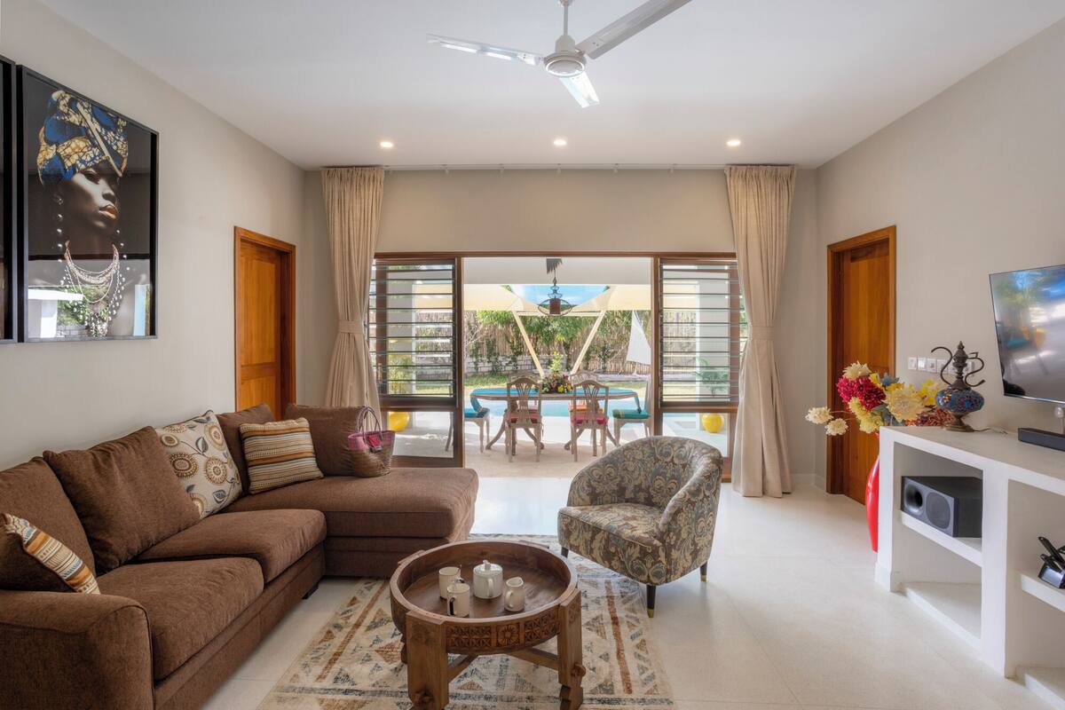 A living room with a brown sofa, patterned cushions, a wooden coffee table, and a patterned armchair. There is a television on a white stand, a vase with flowers, and a ceiling fan.