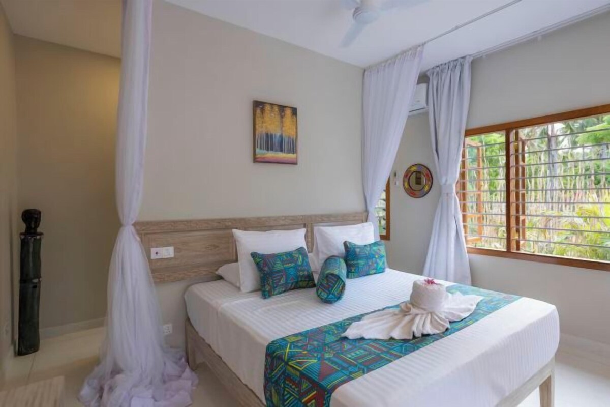 A bedroom with a bed, white curtains, a painting, and a window with a view of greenery.
