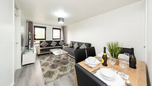 A modern living room with a sofa, a dining table set for two, and a kitchen area in the background.