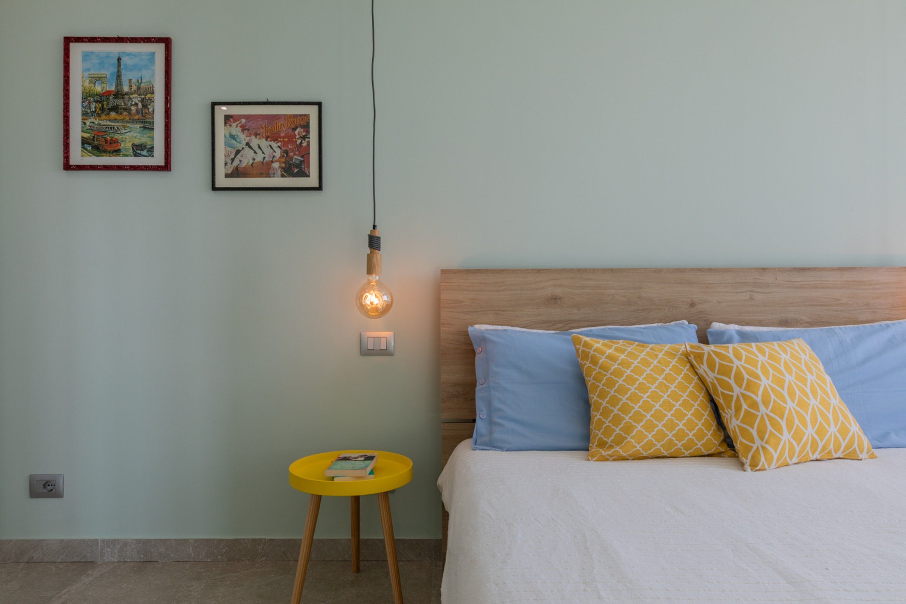 A bedroom with a wooden headboard, a bed with blue and yellow pillows, a small yellow side table with a book, and two framed pictures on the wall.