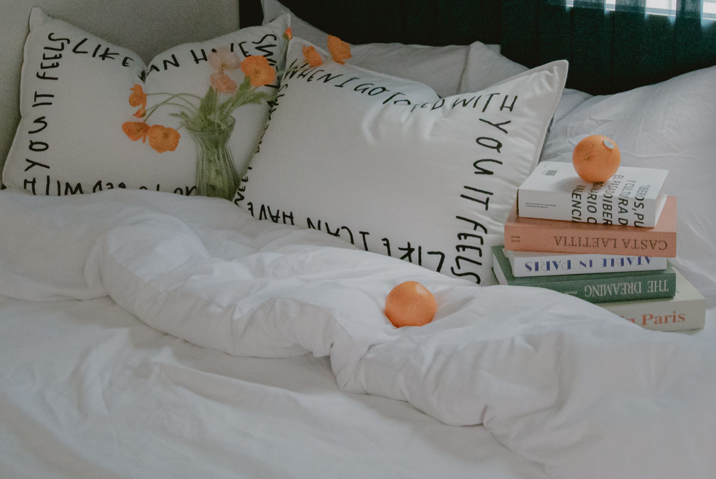 A neatly made bed with white bedding, a vase with flowers, and a stack of books.