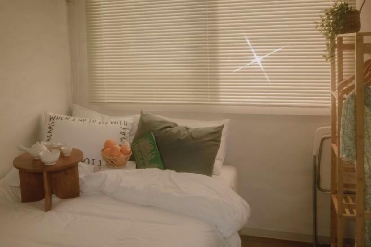 A bedroom with a bed, pillows, a small table with a teapot, and a window with blinds.