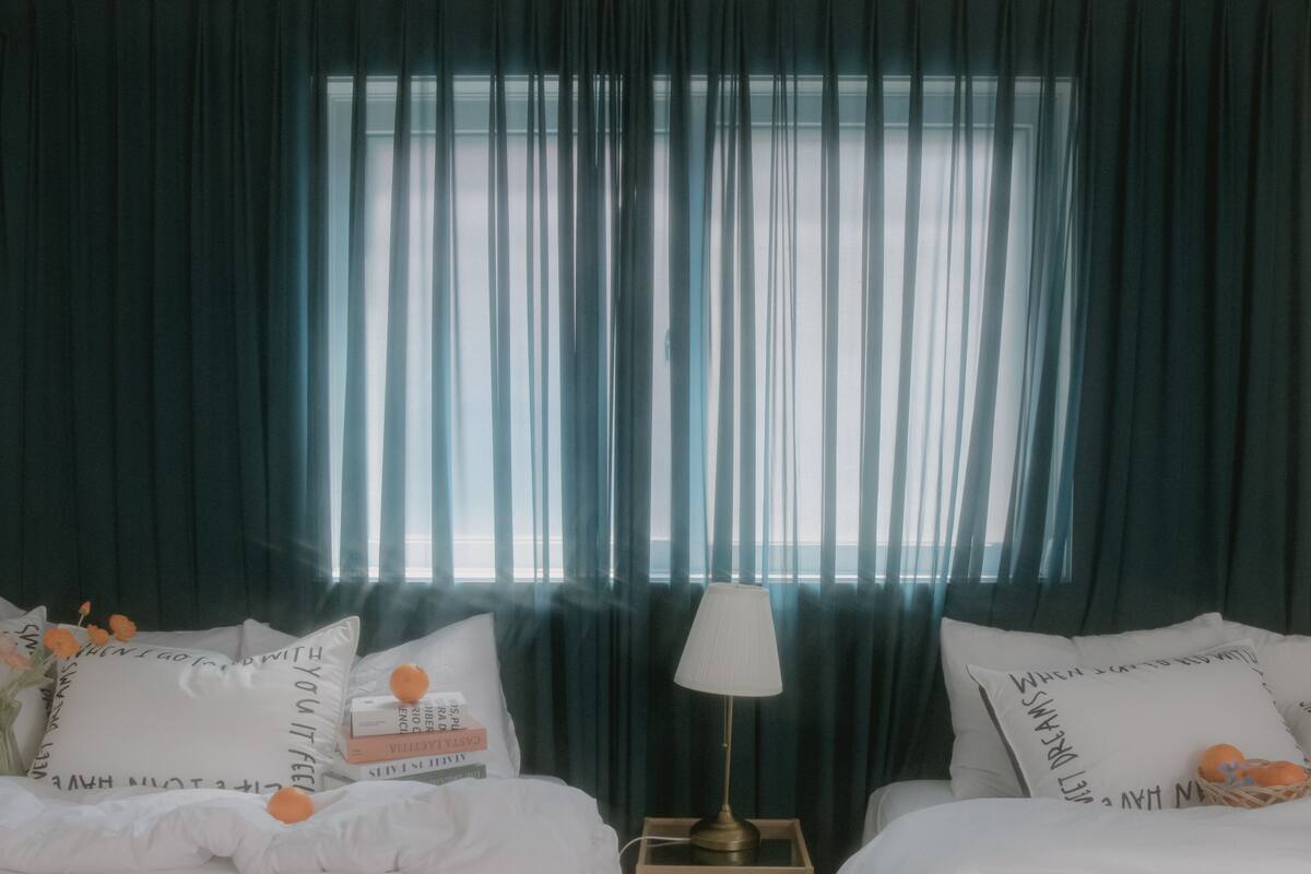 Una camera d'albergo con due letti, un comodino con una lampada e una finestra con tende leggere.