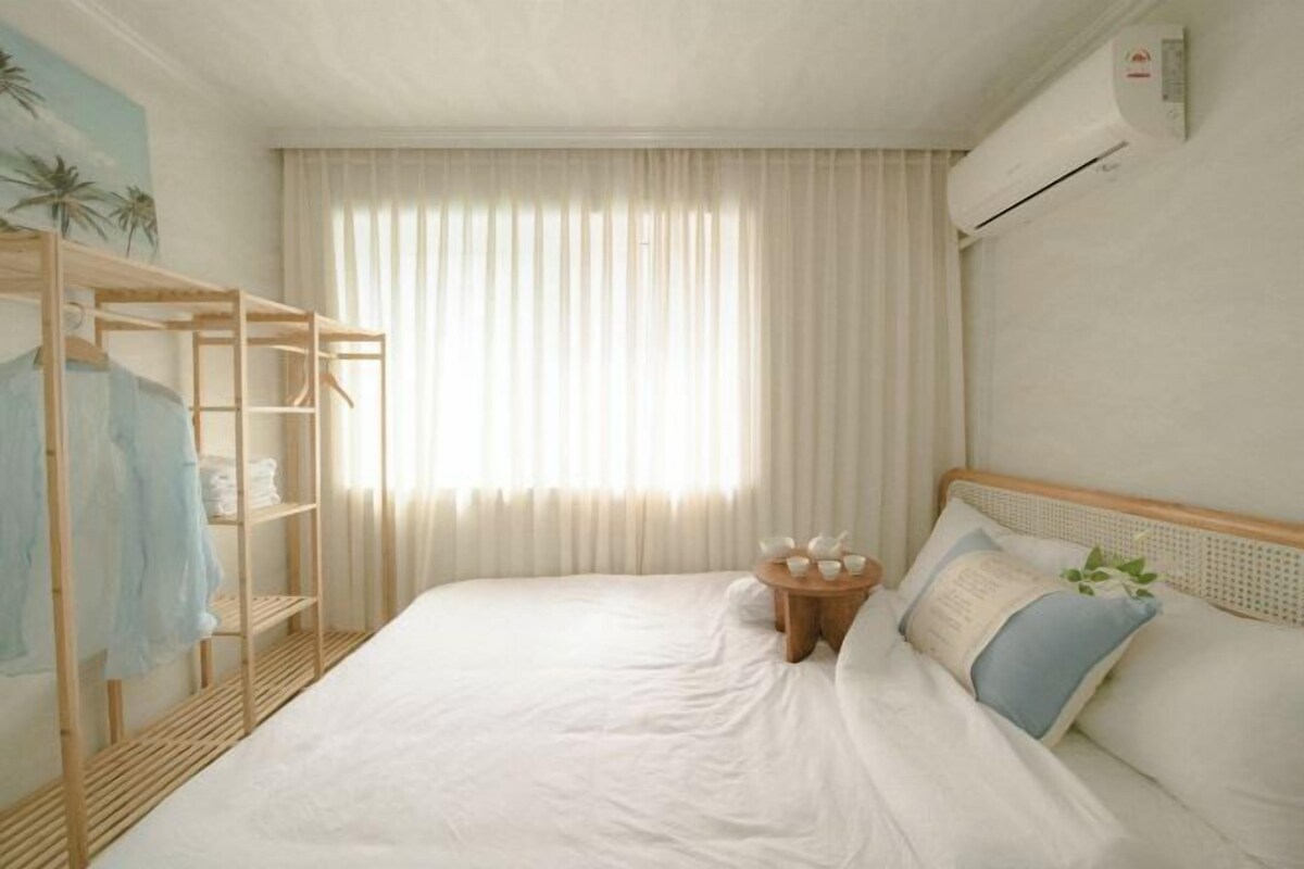A bedroom with a bed, a wooden wardrobe, a small table with cups, and a window with curtains.