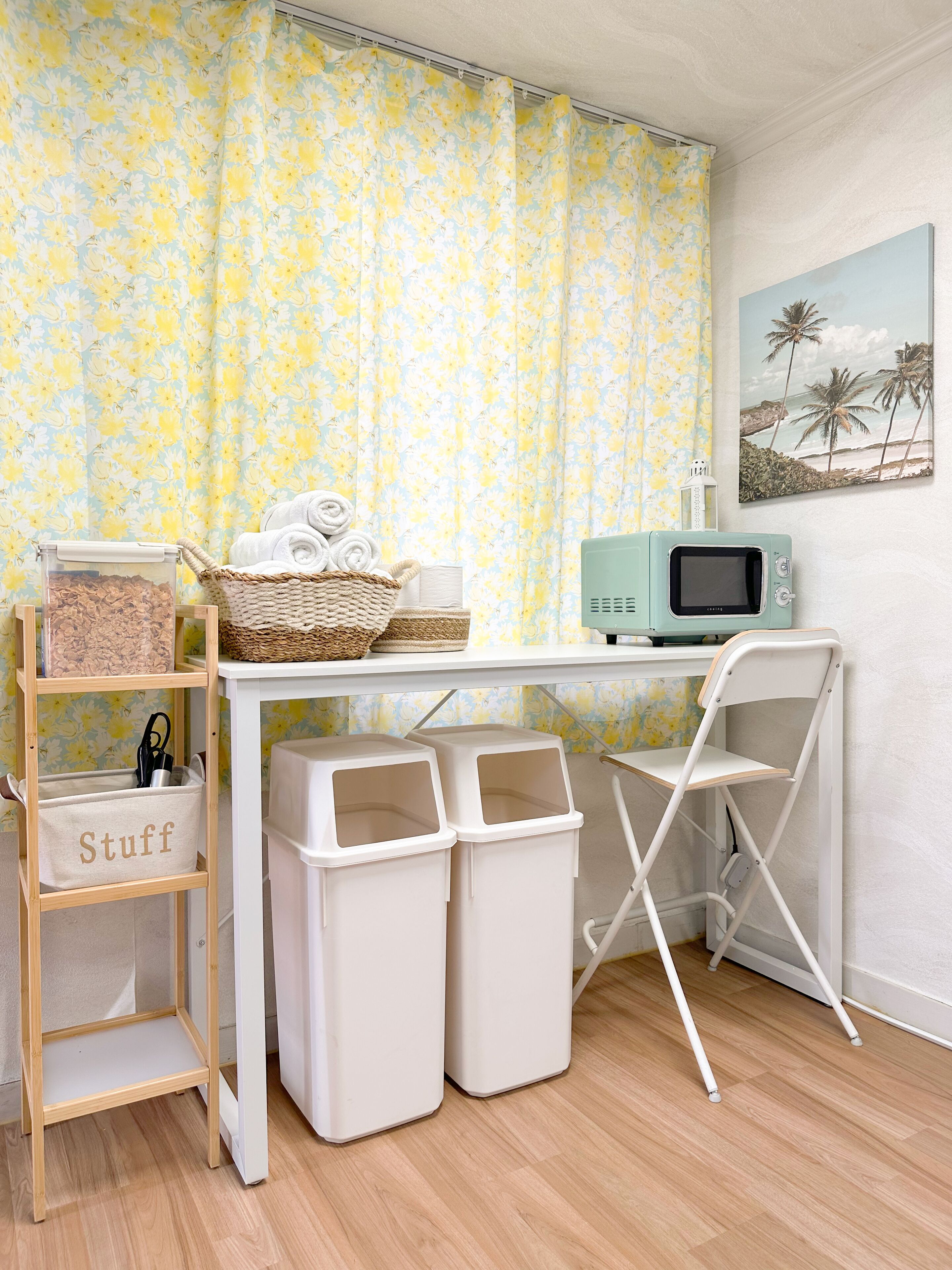 A room with a wooden desk, a microwave, a white chair, two trash bins, a wicker basket with towels, and a shelf labeled "Stuff".