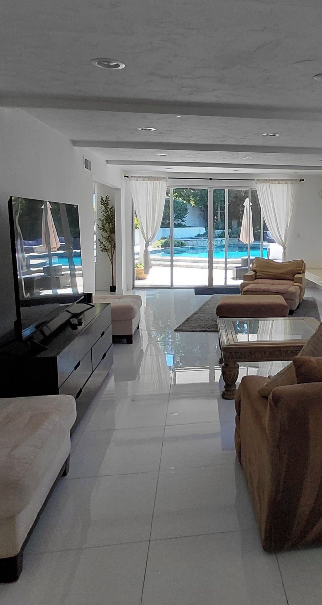 A modern living room with a flat-screen TV, sofas, a coffee table, and a view of a swimming pool through large windows.