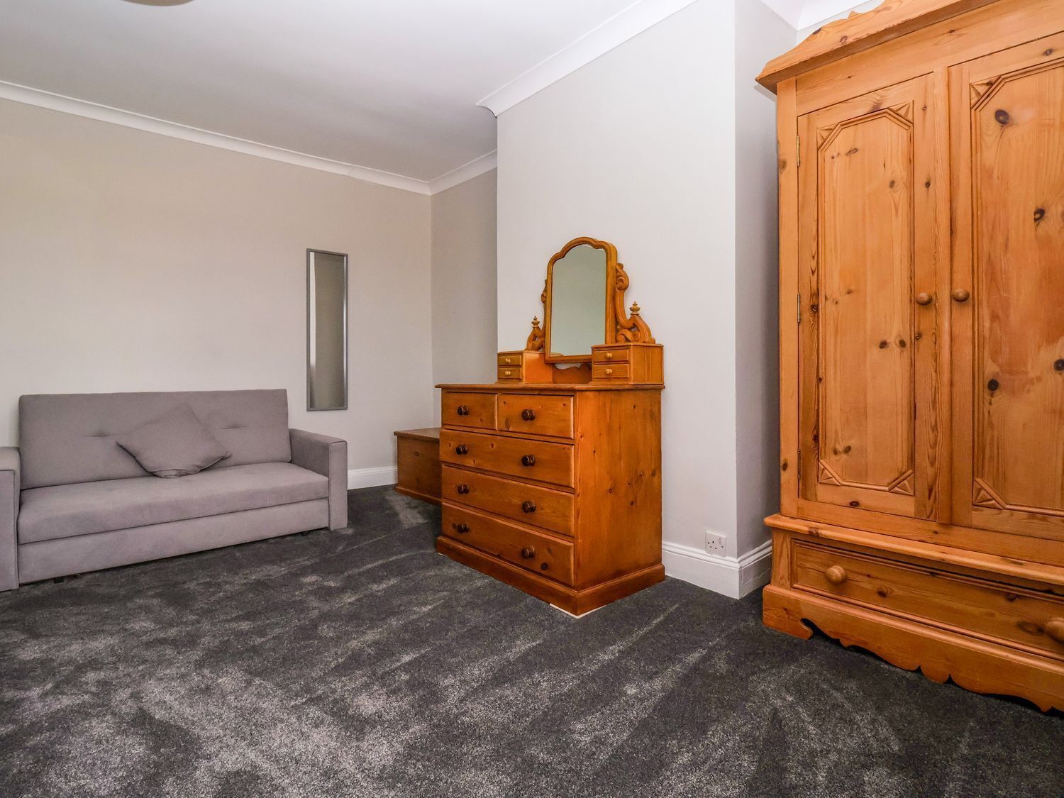 A room with a grey sofa, a wooden dresser with drawers, and a large wooden wardrobe.