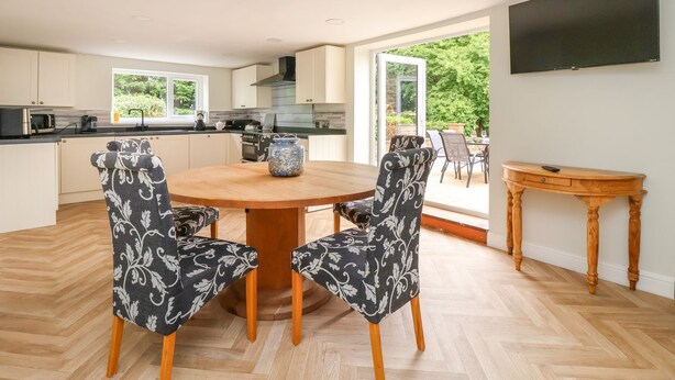 A modern kitchen with white cabinets, a round wooden table, and patterned chairs. A flat-screen TV is mounted on the wall, and a wooden side table is visible. - Bewholme (Driffield)