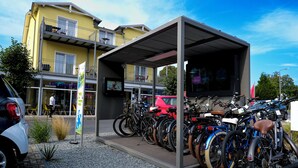 Ein überdachter Fahrradparkplatz mit mehreren Fahrrädern, einem Auto und einem Gebäude mit Außenbestuhlung im Hintergrund.