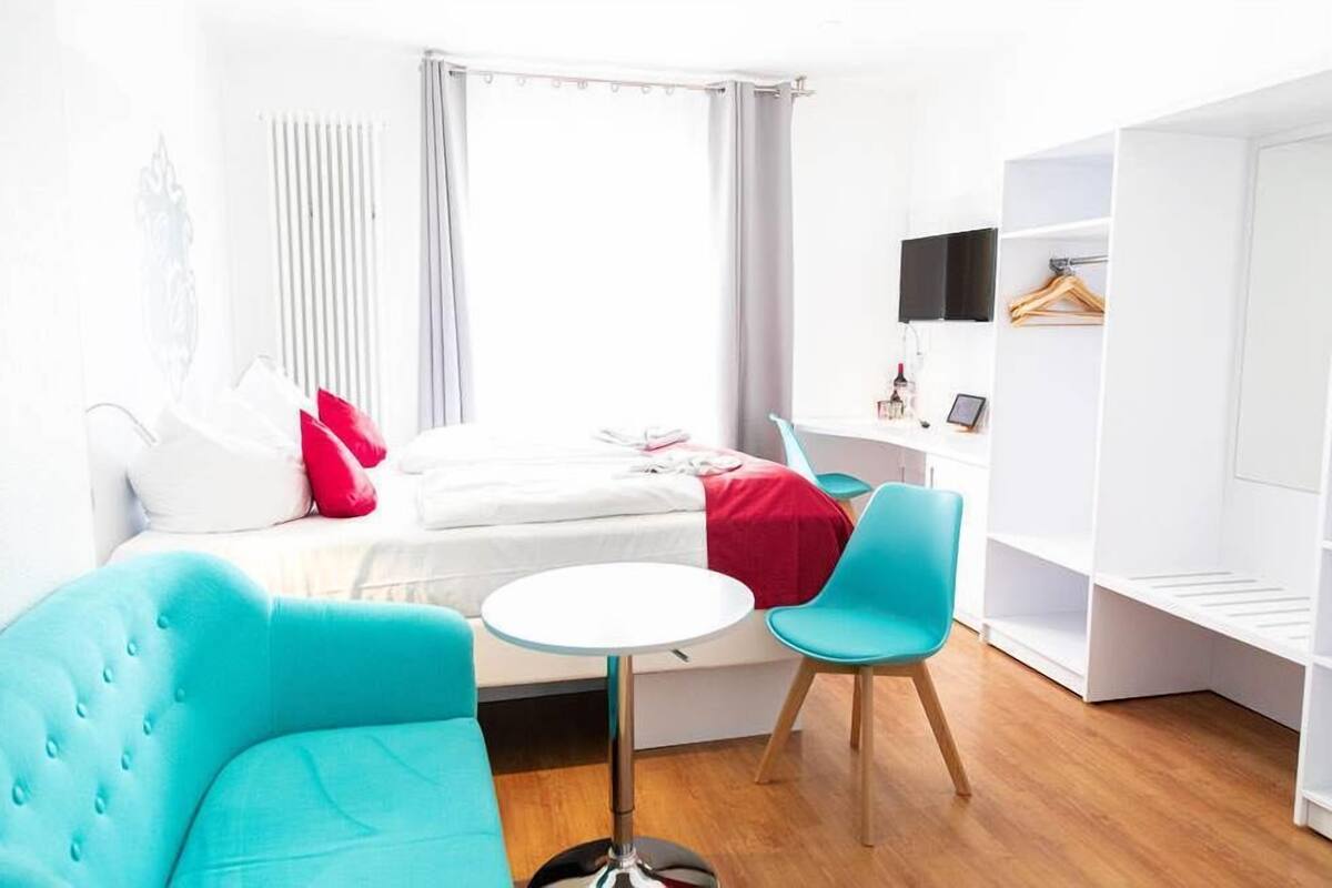 Ein modernes Hotelzimmer mit einem Bett, einem kleinen runden Tisch, zwei Stühlen, einem Kleiderschrank und einem an der Wand montierten Fernseher.