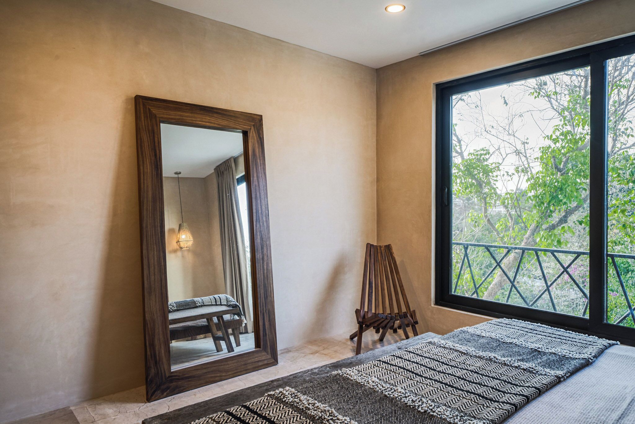 Une chambre avec un grand miroir, un lit recouvert d’une couvre-lit à motifs, une chaise en bois et une fenêtre donnant sur des arbres.