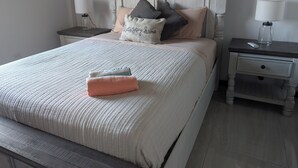 Una cama bien hecha con una colcha estampada y almohadas, un cabecero de madera, y mesitas de noche con lámparas y un tarro de vidrio.