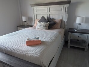 A neatly made bed with a white comforter and pillows, a wooden headboard, two bedside tables with lamps, and a bedside drawer.