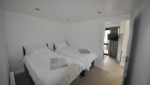 A room with two beds, white bedding, a radiator, and a door leading to another room.