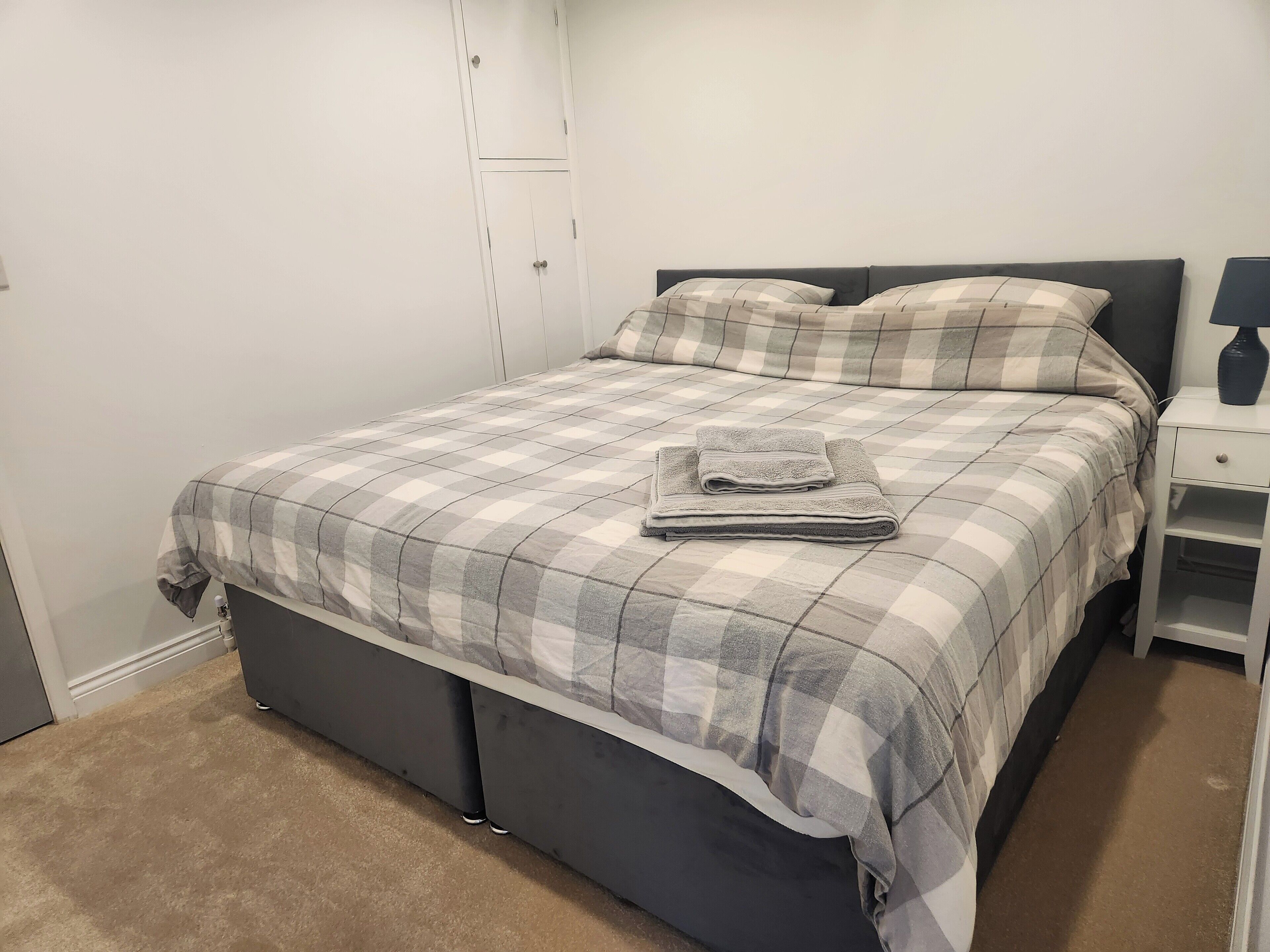 Ein ordentlich bezogenes Bett mit kariertem Überwurf und Kissen, ein Nachttisch mit Lampe sowie ein Schrank an der Wand.