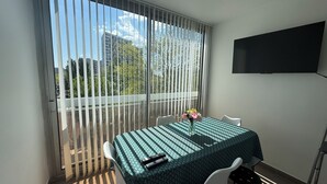 Un balcon avec une table et des chaises, un téléviseur fixé au mur et une vue sur les immeubles et les arbres.