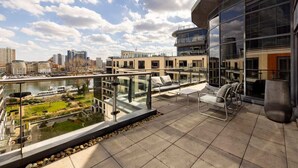 A modern balcony with glass railings, outdoor seating, and a view of a cityscape and river.