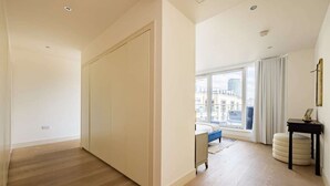 A spacious room with a large wardrobe, a bed with a blue headboard, a small bedside table, and a window with curtains.