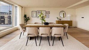 A modern dining room with a large table, six chairs, a vase with flowers, framed artwork, a mirror, and a lamp.