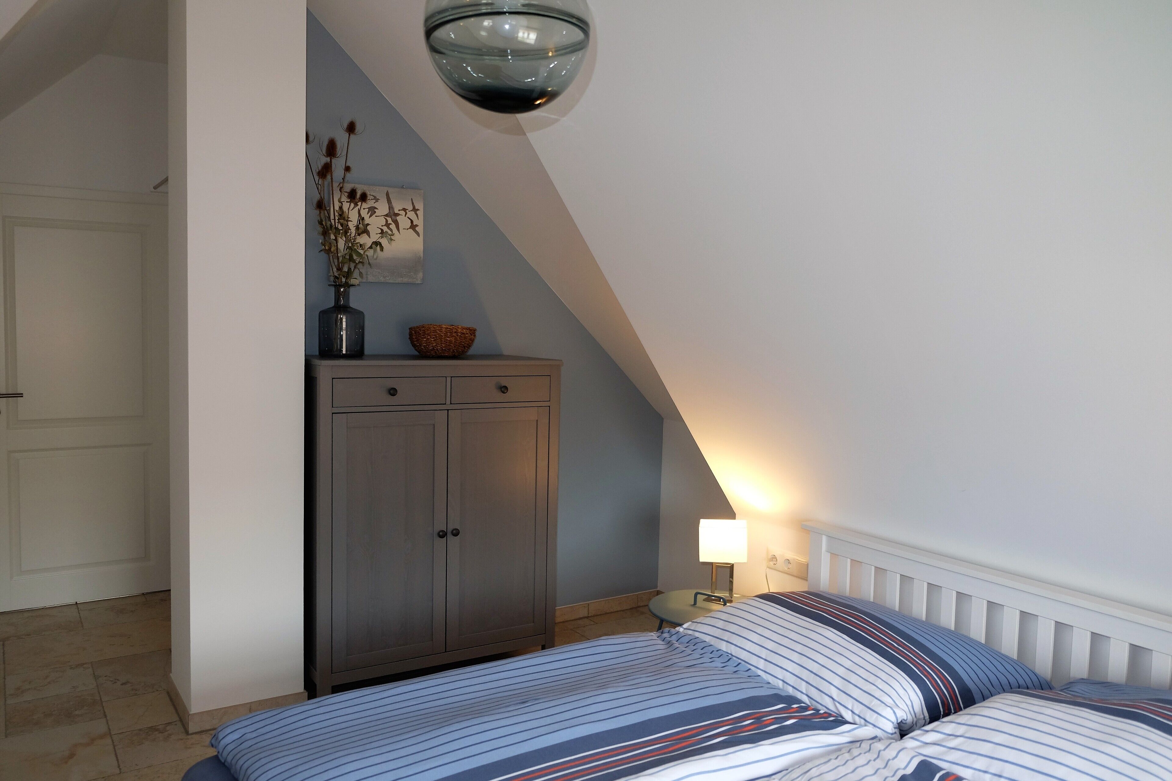 Une chambre à coucher avec un lit, une table de chevet, une lampe, une commode et un vase contenant des fleurs séchées.