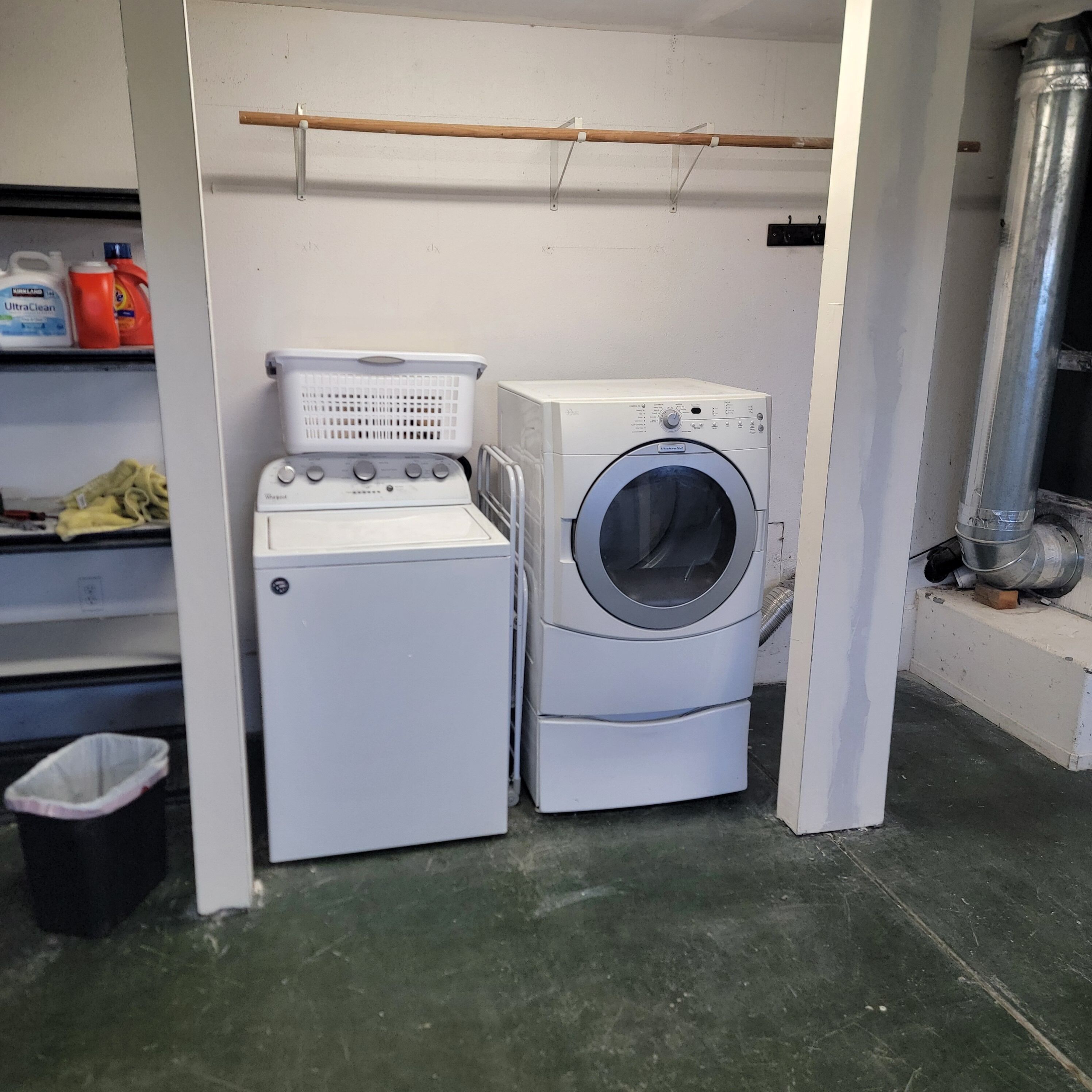 A laundry room with a front-loading washer and dryer, a drying rack, and a shelf with cleaning supplies.