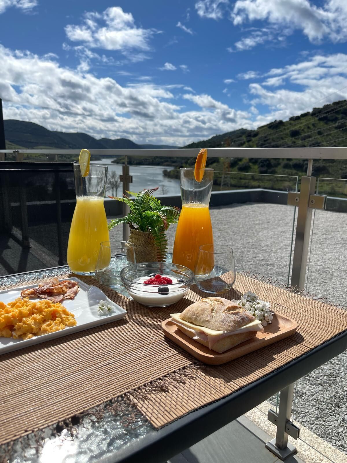 Een ontbijttafel met roerei, toast, yoghurt met bessen en twee sinaasappelsappen op een balkon met uitzicht op een meer.