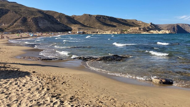 A beach with clear blue water, sandy shore, and rocky hills in the background.