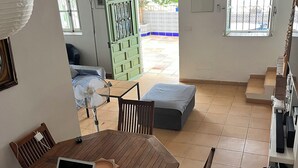 A dining area with a wooden table, chairs, and a laptop. There is a sofa and a window with a view of the outside.