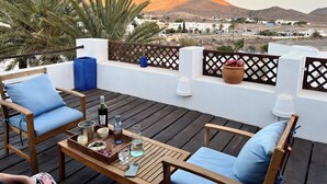 A wooden deck with blue cushioned chairs, a table with drinks and snacks, and a view of a desert landscape with mountains in the background.