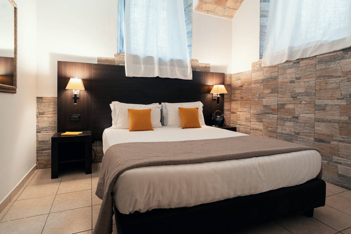 A modern bedroom with a large bed, two bedside tables with lamps, a stone accent wall, and a skylight.