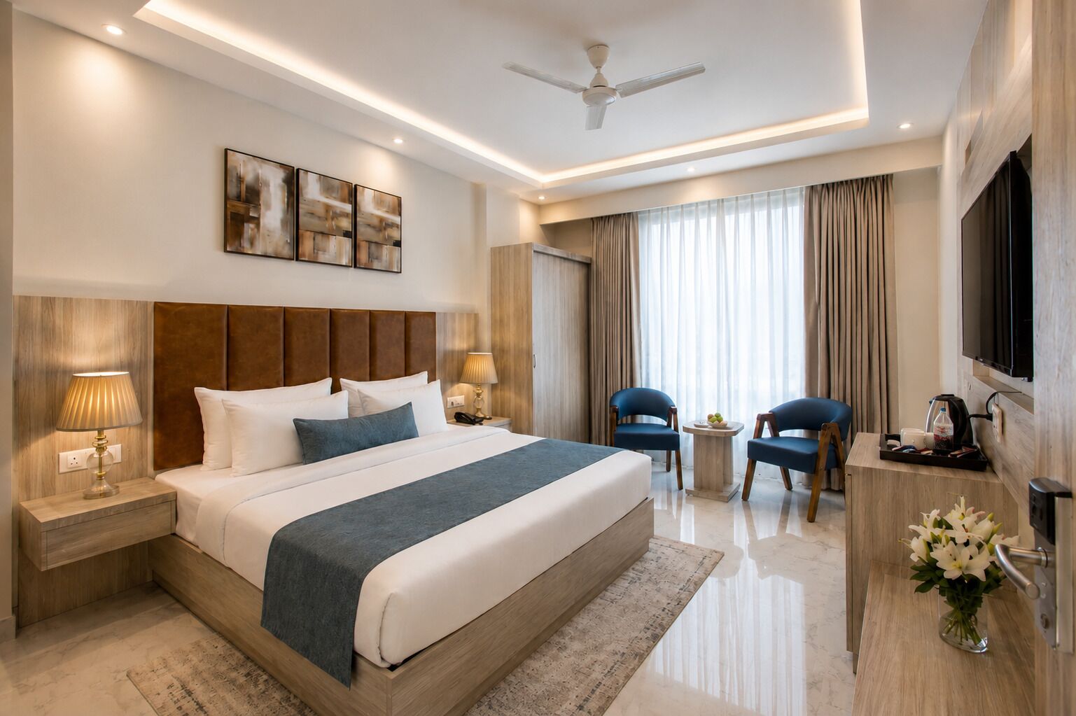 A modern hotel room with a large bed, bedside tables, a desk, chairs, a TV, and artwork on the wall.