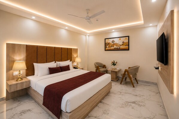 A modern hotel room with a large bed, bedside lamps, a ceiling fan, a small table with a vase, two armchairs, and a flat-screen TV mounted on the wall. - Hotel West End Mahipalpur Delhi IGI (New Delhi)