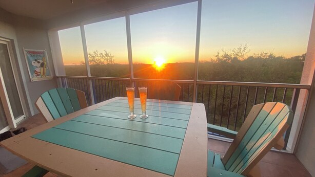 A balcony with a table and chairs, two glasses of a beverage, and a view of the sunset.