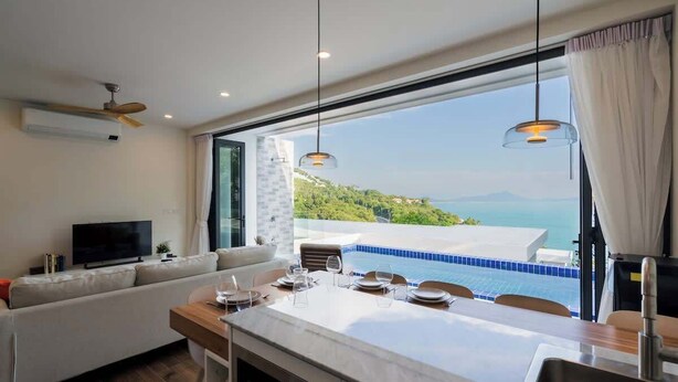 A modern kitchen with a large island, a dining area with a view of the sea, and a living area with a sofa and TV.
