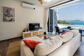 A modern living room with a flat-screen TV, a sofa with cushions, a coffee table, and a view of the sea through a glass door.