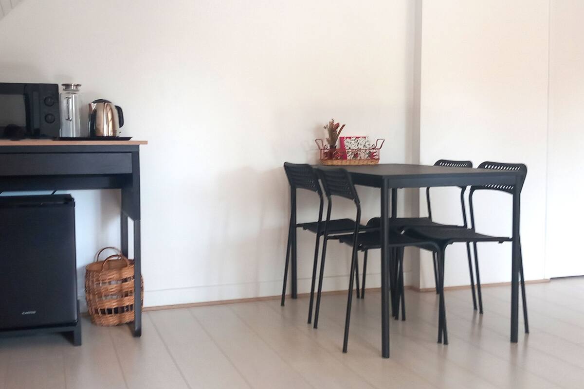 Eine kompakte Küche mit Mikrowelle, Wasserkocher und kleinem Kühlschrank. Ein Esstisch mit vier Stühlen und ein Korb auf der Arbeitsfläche.