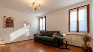 A spacious living room with a dark green sofa, a chandelier, a radiator, and a window with blinds.